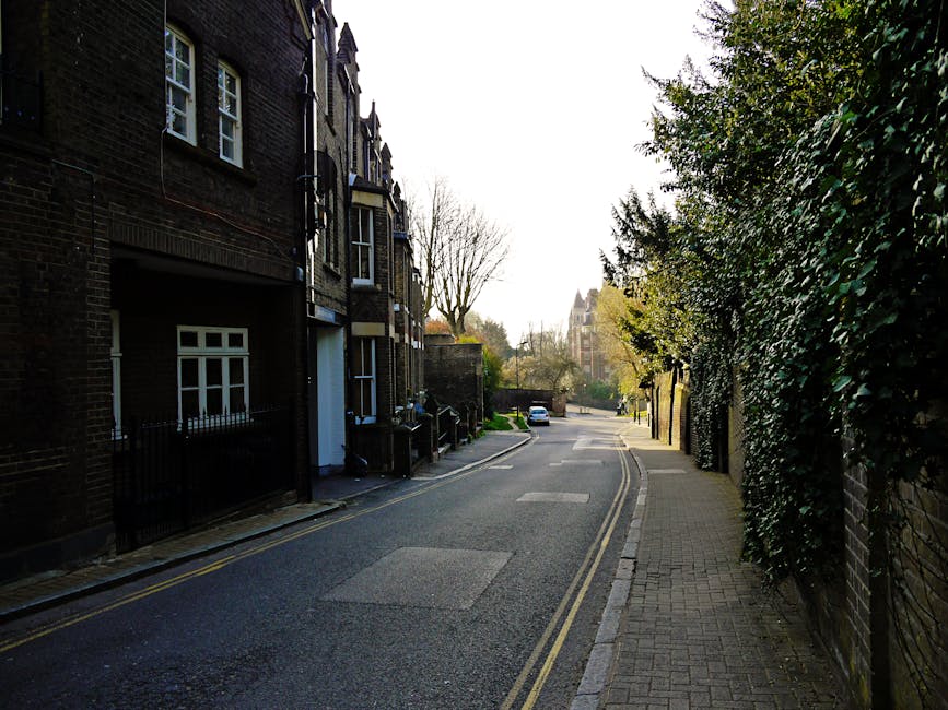 A narrow, winding dirt pathway in a park or garden setting, surrounded by lush green trees with dense foliage. A weathered wooden bench is positioned on the left side of the path, partially shaded by 