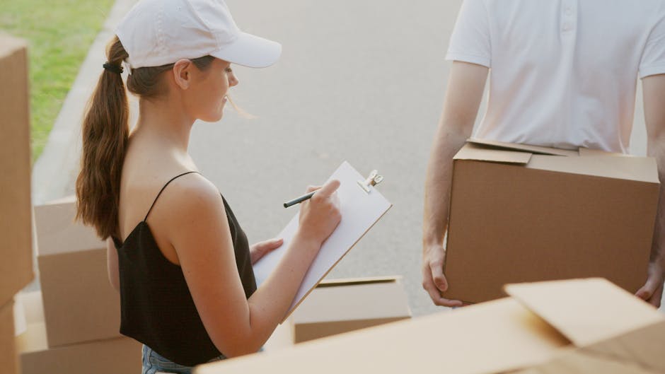 A man and a young girl are inside a home during the packing and moving process. The man, with dark hair tied back, is leaning over a large cardboard box, observing as the girl, with curly hair, holds 